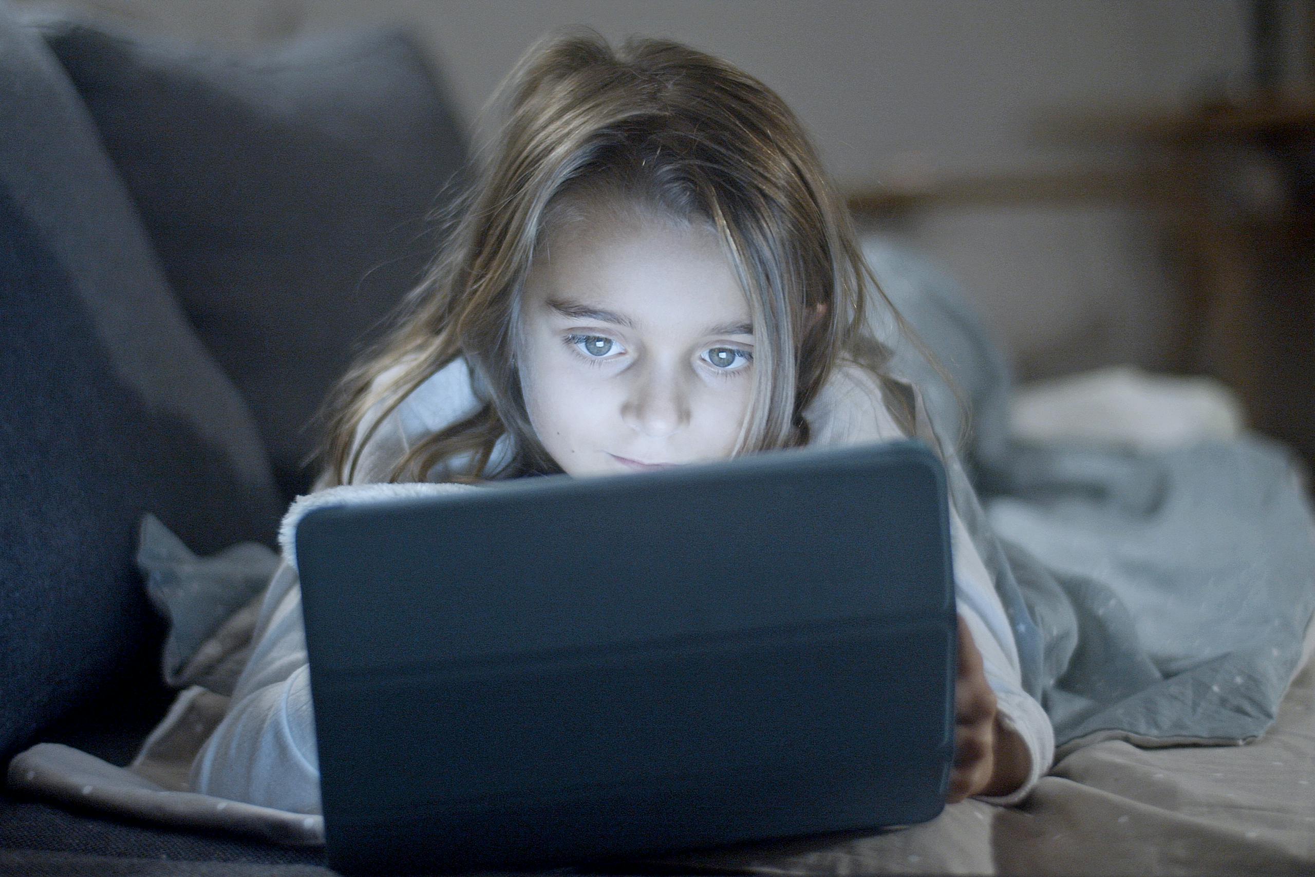 A young girl with blond hair uses a tablet in bed, focused and engaged.